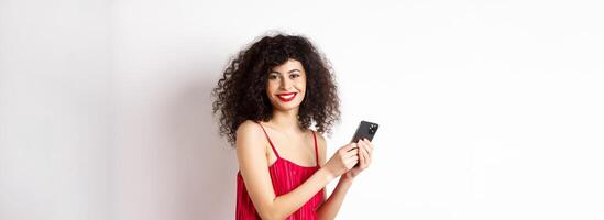 Happy elegant woman in red dress writing message, using smartphone and smiling at camera, standing against white background photo