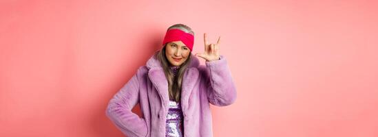 Funny and cool asian senior woman showing rock n roll gesture, looking sassy, standing over pink background photo