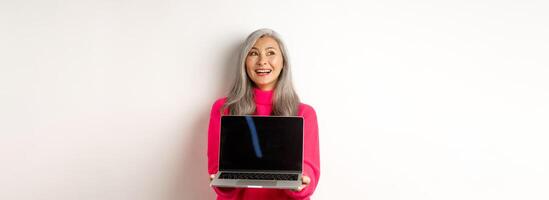 E-commerce concept. Happy beautiful asian senior woman with grey hair, looking aside and smiling, showing laptop black screen, standing over white background photo