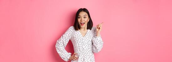 Cute asian woman in dress pointing upper left corner copy space. Korean model showing advertisement, standing over pink background photo