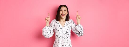 Amazed brunette girl in dress looking, pointing fingers up, showing logo with wondered smile, standing over pink background photo