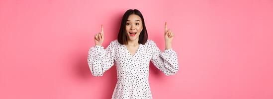 Amazed brunette asian girl pointing fingers up, showing advertisement, demonstrate spring promo offer, standing over pink background photo