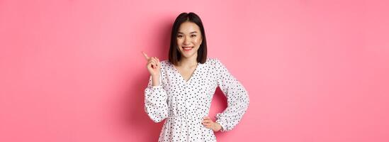 Beautiful asian female model smiling, pointing finger at upper right corner copy space, showing advertisement banner, standing over pink background photo
