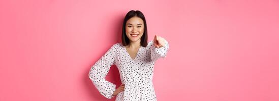 Confident asian woman in dress pointing at you, smiling and staring at camera, standing against pink background photo