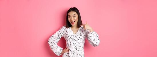 Beautiful asian girl in dress looking happy, pointing finger at herself, standing on romantic pink background photo