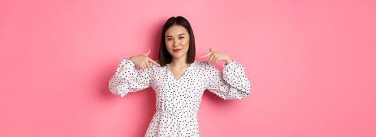 Beautiful asian woman looking confident, pointing at herself and smiling, showing logo on chest, standing over pink background photo
