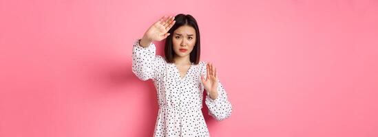 Timid and scared asian woman defending herself, raising arms in protection, victim being attacked, standing over pink background photo