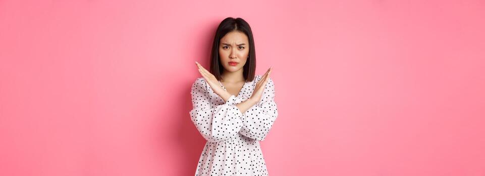 enojado asiático mujer contar detener, haciendo cruzar a prohibir o desaprobar algo, ceñudo disgustado, en pie terminado rosado antecedentes foto