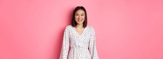 Happy korean woman in dress looking at camera, smiling and laughing with sincere expression, standing over pink background photo