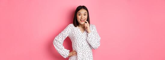 Image of beautiful young lady in dress, biting finger and smiling, staring with temptation and amazement, want buy something, standing over pink background photo