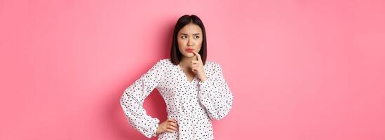 Troubled asian girl making decision, frowning and touching lip while thinking, looking unsure at upper left corner and choosing, standing over pink background photo