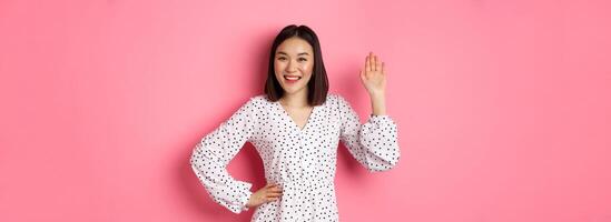 Beautiful asian woman in dress saying hello, waving hand to greet and say hi, smiling friendly at camera, standing over pink background photo