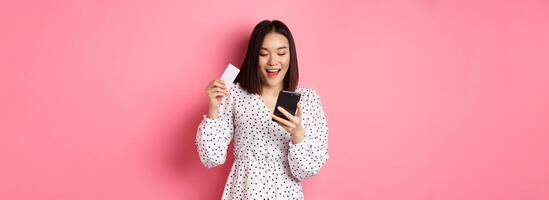 Attractive young asian woman order online, holding credit card and mobile phone, making internet purchase, standing happy over pink background photo