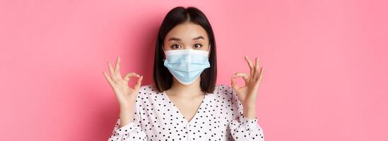 Covid-19, pandemic and lifestyle concept. Image of cute asian woman in face mask showing its okay gesture, make ok signs, have all under control, standing over pink background photo