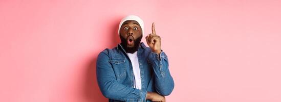 Handsome hipster Black guy having an idea, raising finger up and saying suggestion, got solution, standing over pink background photo