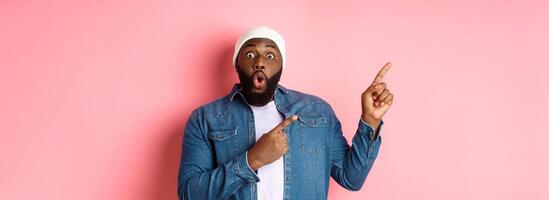 Amazed Black guy showing awesome promo offer, staring at camera excited and smiling, pointing fingers at upper left corner banner, pink background photo