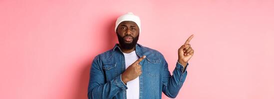 Skeptical and doubtful Black man staring at camera unsure, pointing fingers upper left corner at something uncertain, standing over pink background photo