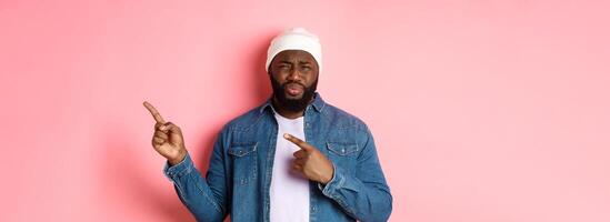 Disappointed and skeptical african-american guy complaining, cringe from disgust, frowning and grimacing from dissatisfaction, pointing fingers right, pink background photo