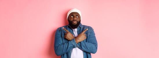 Confused Black man with beard, making choice, pointing fingers sideways and looking puzzled, standing over pink background photo