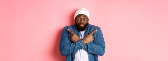Confused Black man asking help with choice, pointing sideways and staring at camera, making decision, standing over pink background photo