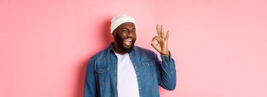 Happy smiling african american man showing okay sign, approve and praise good offer, standing over pink background photo