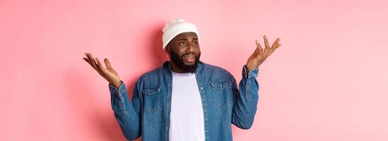 Skeptical confused man looking at something riddiculous, staring left and shrugging, raising hands up puzzled, standing over pink background photo
