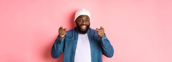 Happy african-american hipster guy pointing at camera, need you, smiling and inviting to company, standing over pink background photo