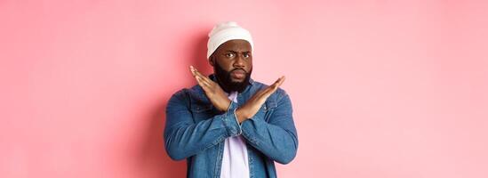 Disappointed Black man, staring at camera displeased and frowning, showing stop cross gesture, prohibit and disagree, standing over pink background photo