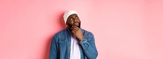 Image of puzzled and concerned black man, staring at upper left corner and thinking, making decision with doubts, standing over pink background photo