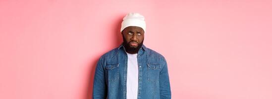 Disappointed and sad Black man, sulking and whining, looking at upper left corner with offended and jealous grimace, standing over pink background photo