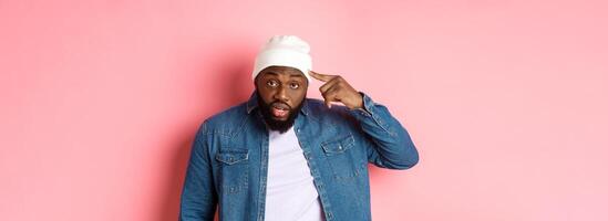 Annoyed and pissed-off african-american man pointing finger at head, scolding someone stupid, staring bothered at camera, pink background photo