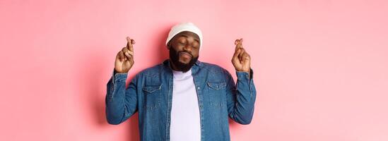 Hopeful and dreamy Black man making wish, cross fingers and smiling, standing over pink background photo