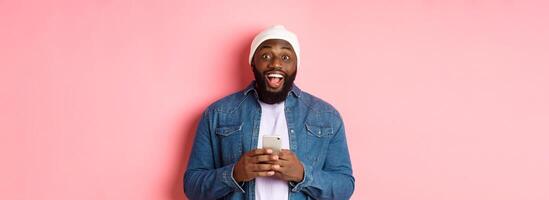 Technology and online shopping concept. Surprised young Black man using mobile phone, looking at camera amazed and happy after reading message, pink background photo