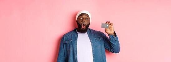 Shopping concept. Amazed Black guy showing credit card, staring at camera impressed, recommending bank, standing over pink background photo