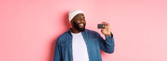 Shopping concept. Image of happy african-american man looking satisfied at credit card, recommending bank, standing over pink background photo