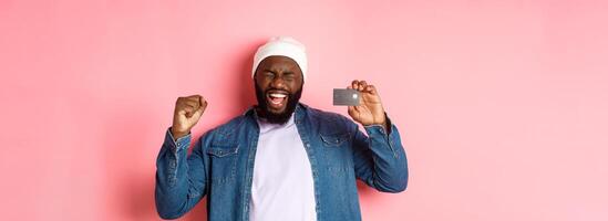 Shopping concept. Happy Black man rejoicing, scream of joy and showing credit card, standing over pink background photo