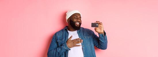 Shopping concept. Image of happy african-american man looking satisfied at credit card, recommending bank, standing over pink background photo