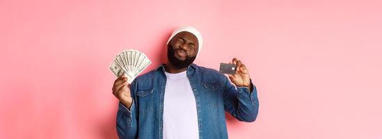 Shopping concept. Sad and whining Black guy showing credit card and dollars, grimacing reluctant, standing over pink background photo