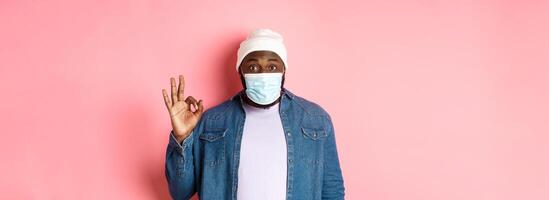Coronavirus, lifestyle and social distancing concept. Impressed african-american man in face mask, showing okay sign, like and agree, standing over pink background photo