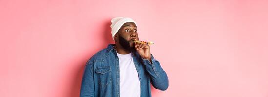Funny Black man blowing in party whistle and pouting, staring sideways with popped eyes, standing against pink background photo