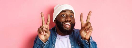 Close-up of cheerful african-american hipster in beanie smiling, showing peace signs, standing happy against pink background photo