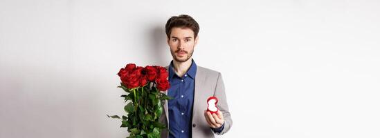 Handsome man in suit going to make a proposal, standing with red roses flowers and engagement rin in box, making romantic surprise, white background photo