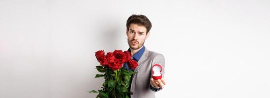 Handsome boyfriend in suit asking to marry him, standing with red bouquet of roses and engagement ring, looking pleading at camera, standing over white background photo