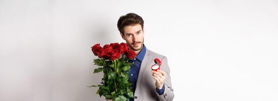 Handsome man in suit, showing engagement ring and looking romantic at camera, standing with red roses over white background. Valentines day and love concept photo