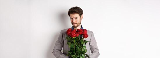 Indecisive bearded man in suit, holding flowers roses and looking at camera unsure, going on romantic date, standing over white background photo