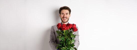 Handsome man in love wishing happy valentines day, giving bouquet of flowers on romantic date, smiling at camera, wearing suit over white background photo