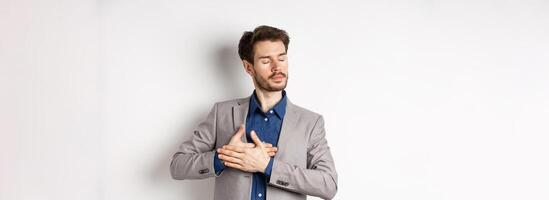 Romantic bearded man in stylish suit holding hands on heart, close eyes and remember something with tender feelings, standing nostalgic on white background photo