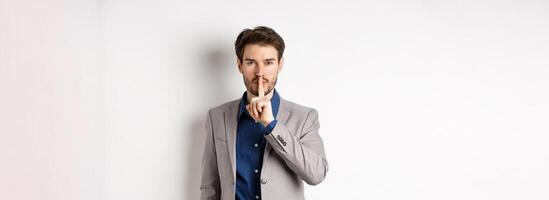 Handsome macho man in suit shushing at camera, press finger to lips, ask keep voice down, tell secret, standing against white background photo