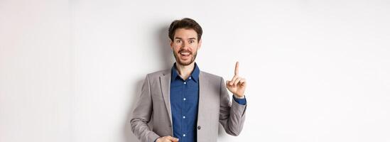Excited businessman in suit pitching good idea, raising finger in eureka sign and looking amused, saying suggestion, standing on white background photo