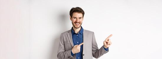 Handsome caucasian man in business suit pointing fingers left at banner and smiling, showing advertisement, standing against white background photo